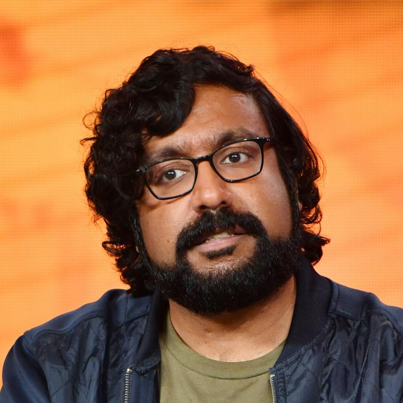 Comedian Hari Kondabolu listens on stage against an orange backdrop